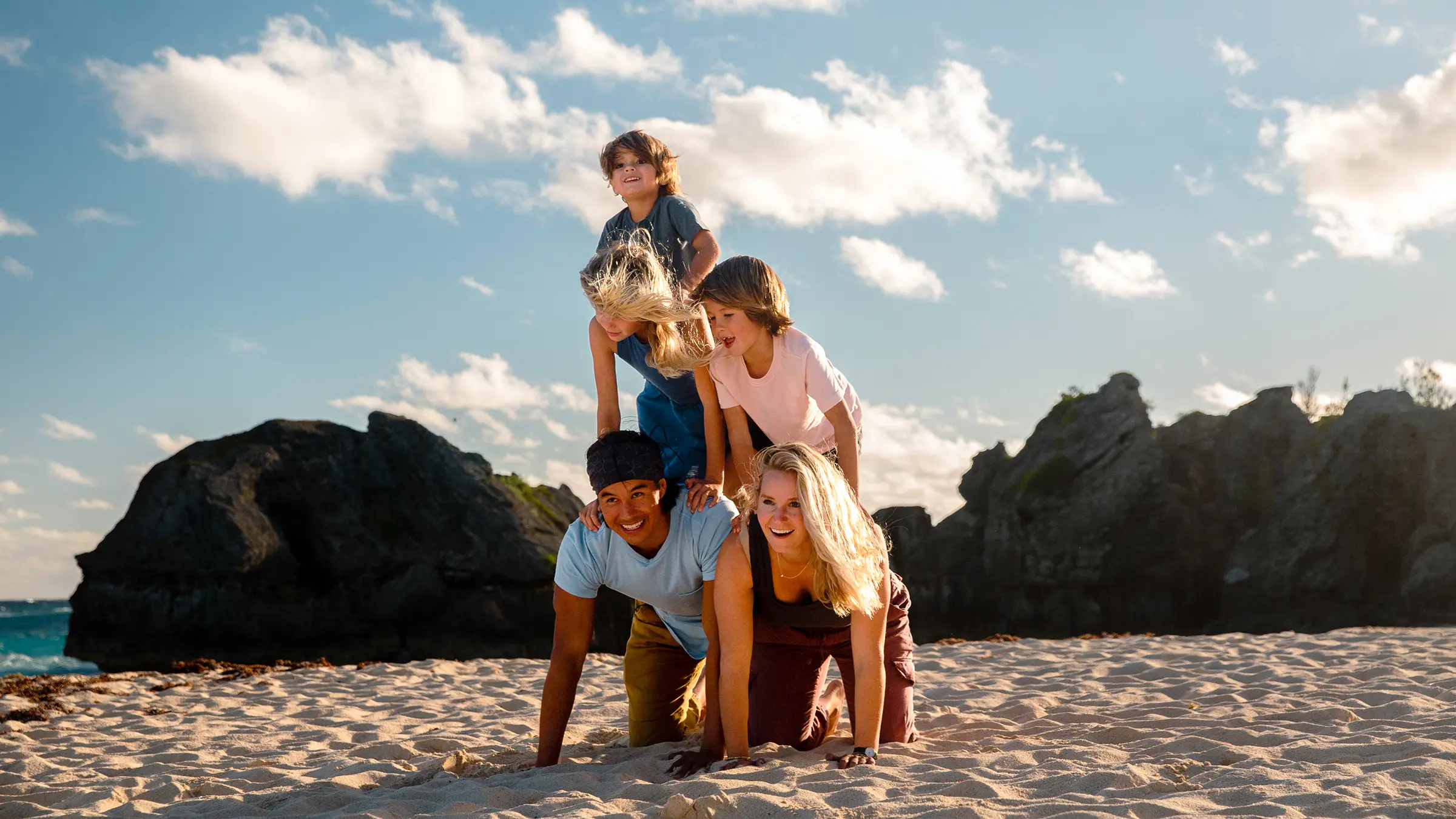 Family stacked playfully on a sandy beach beside dark rocks